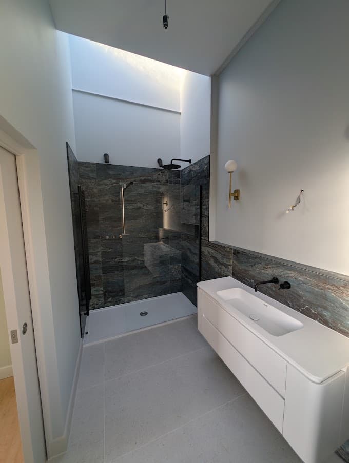 A finished bathroom with dark stone-tiled shower walls, a white floating vanity and painted walls and ceiling under a skylight