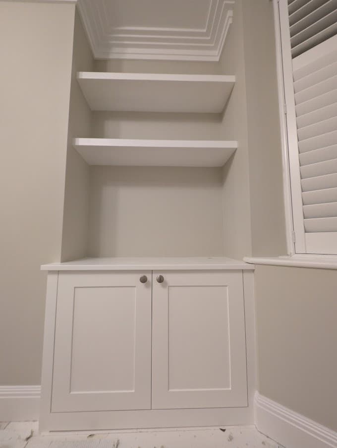 Painted built-in alcove shelving and a base cupboard finished in white, beside a shuttered window