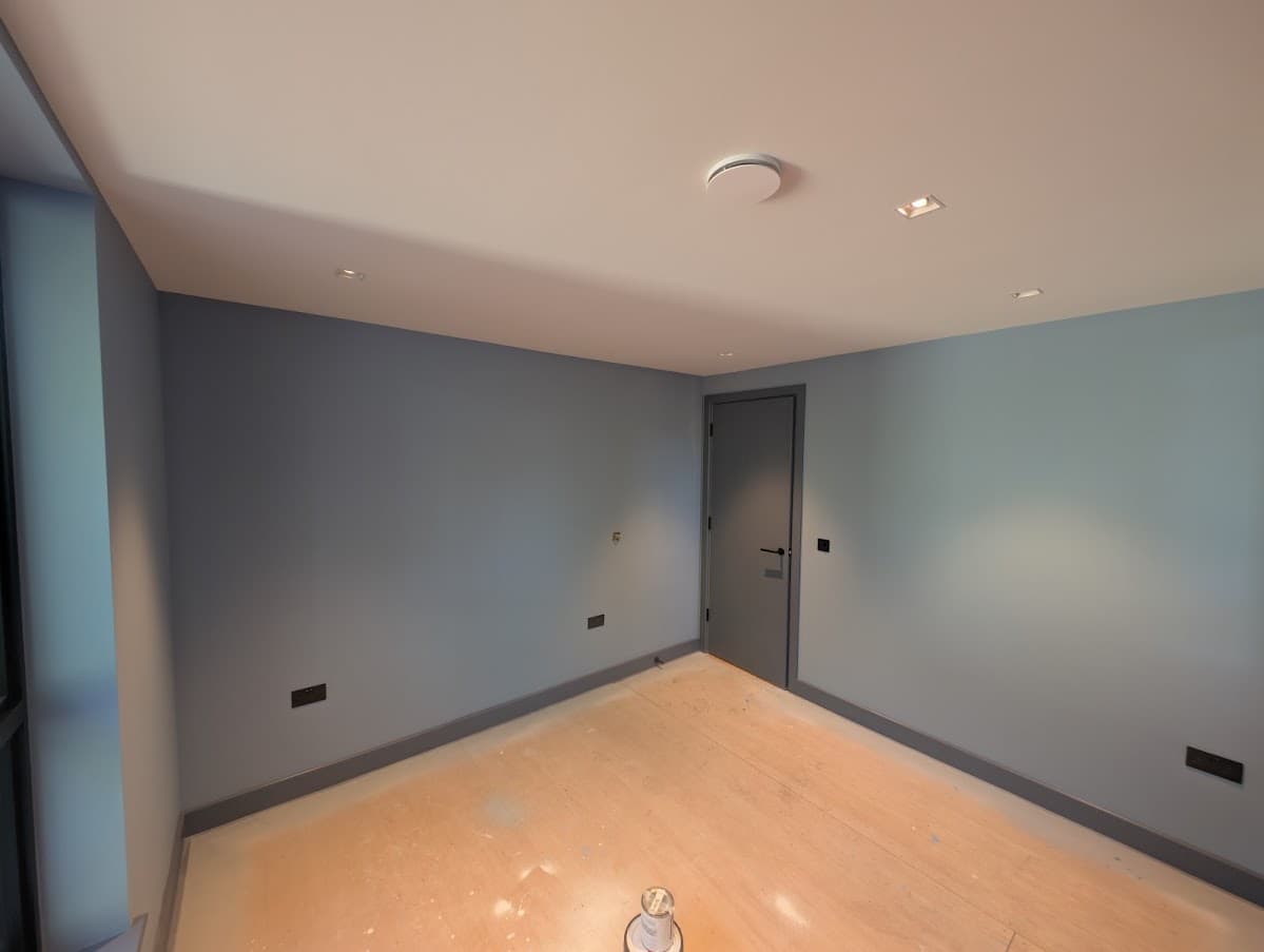 An empty room painted in a soft blue-grey, with a fitted door and recessed ceiling lighting, awaiting flooring