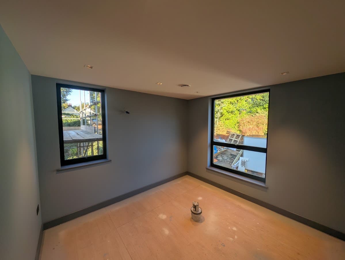 An empty room with sage green walls, bare floorboards and two black-framed windows overlooking neighbouring gardens and rooftops
