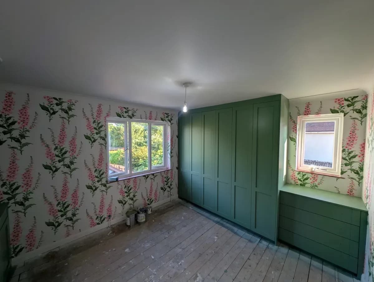 Bedroom with floor-to-ceiling fitted wardrobes and a low drawer unit finished in deep green, beside walls hung with a pink and green floral wallpaper