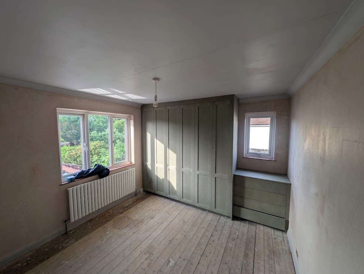 Fitted wardrobes and a low drawer unit sprayed sage green in a bedroom with bare floorboards and two windows