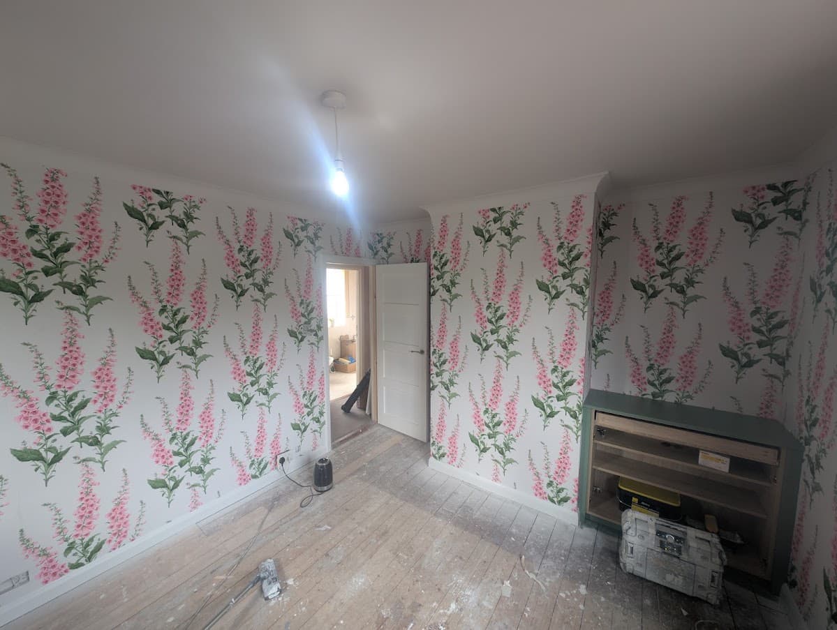 A bedroom with pink and green foxglove-pattern wallpaper hung across two walls and an open doorway between them