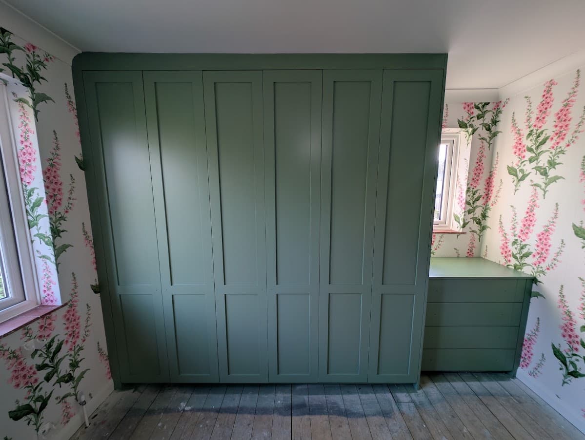 Finished sage green fitted wardrobes flanked by pink foxglove-pattern wallpaper, with a matching green window seat and drawer unit