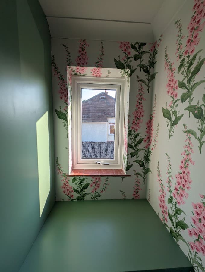 A built-in window nook with pink foxglove-pattern wallpaper on three walls and a sage green painted bench seat