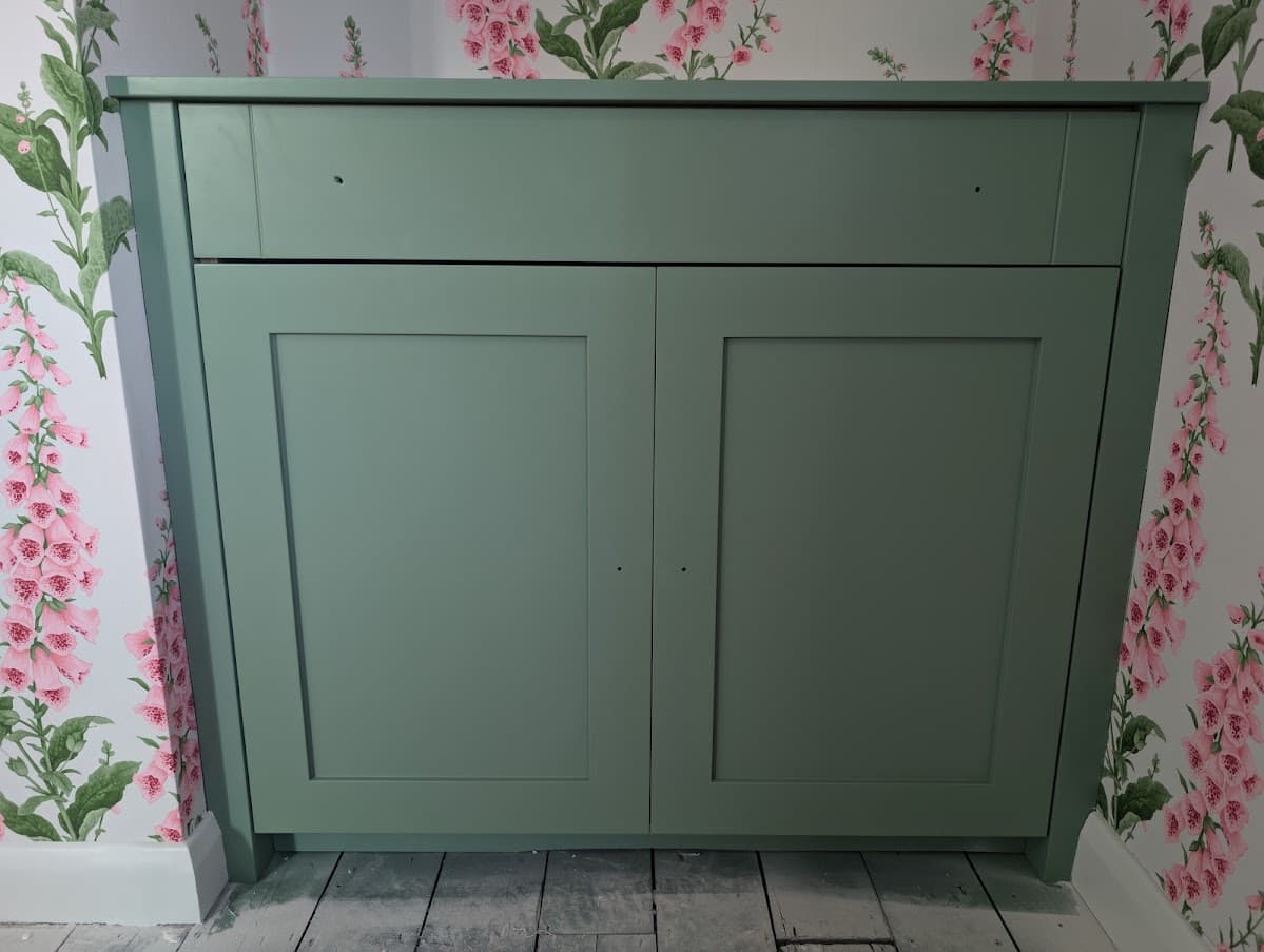Close view of a sage green built-in cabinet with two shaker-style doors, set against pink foxglove-pattern wallpaper