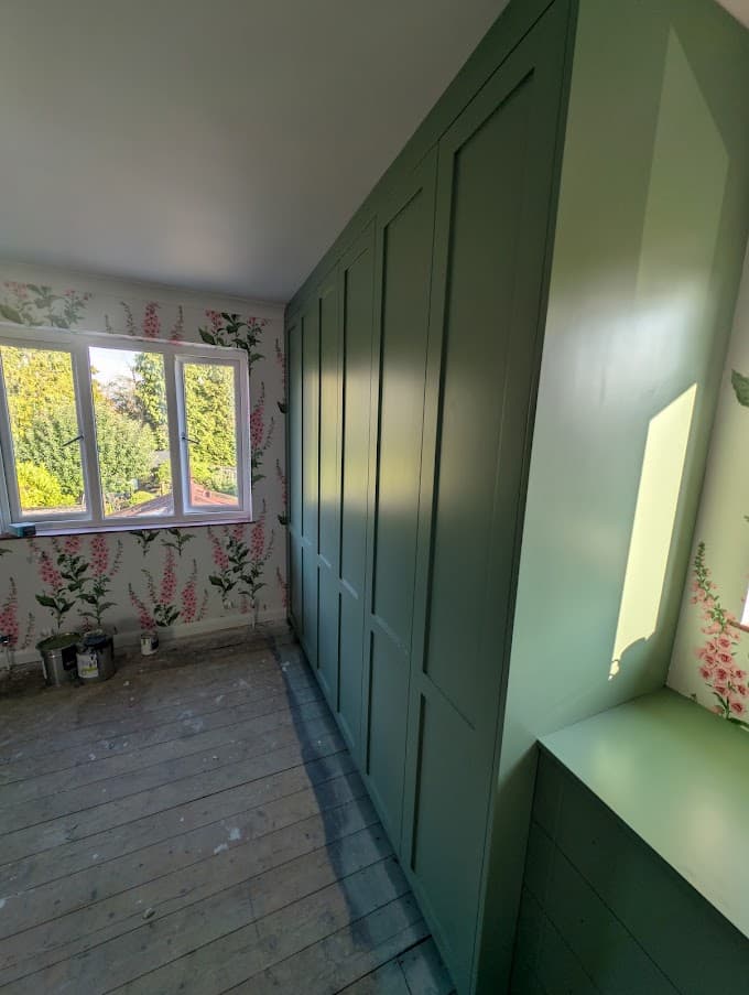 Sage green fitted wardrobes meeting a built-in window bay bench, with pink foxglove-pattern wallpaper on the surrounding walls