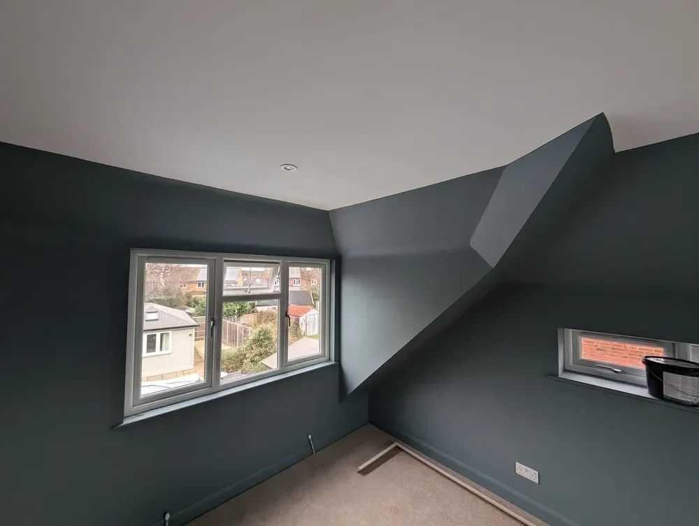 Loft bedroom with angled walls and dormer returns painted in a deep slate colour, meeting a white ceiling above a white-framed window