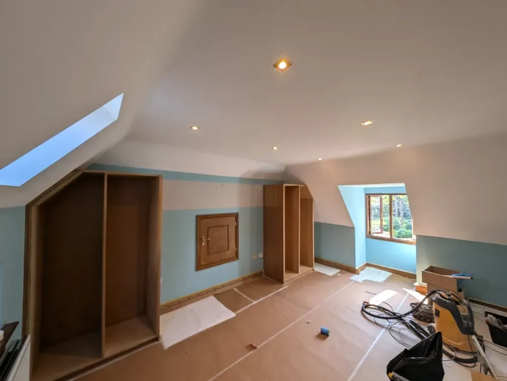 The same loft bedroom mid-job: floors covered with protective sheeting, a sander and equipment on site, bare timber wardrobe carcasses and part-painted walls