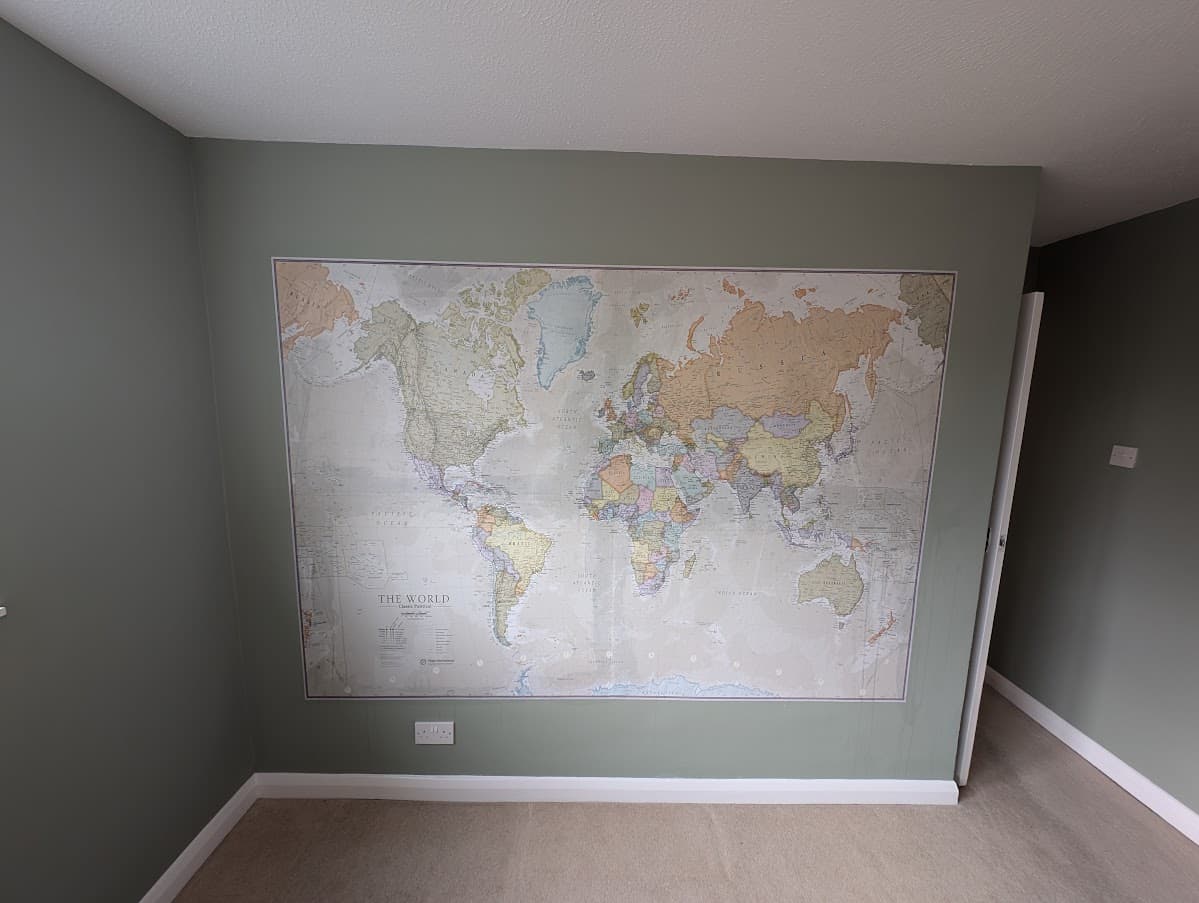 A sage green bedroom wall with a large framed world map wallpaper mural, fully hung and finished