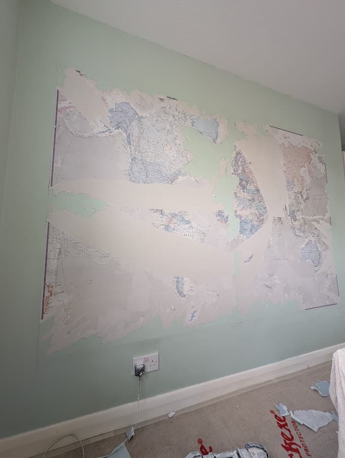The same wall with the old world map wallpaper part-stripped, torn paper hanging from a sage green wall above a skirting board