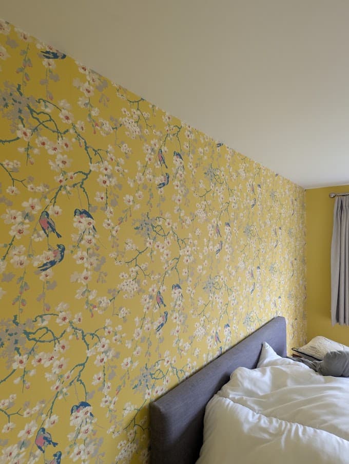 A bedroom with yellow botanical wallpaper patterned with birds and blossom, covering a sloped ceiling wall above a bed